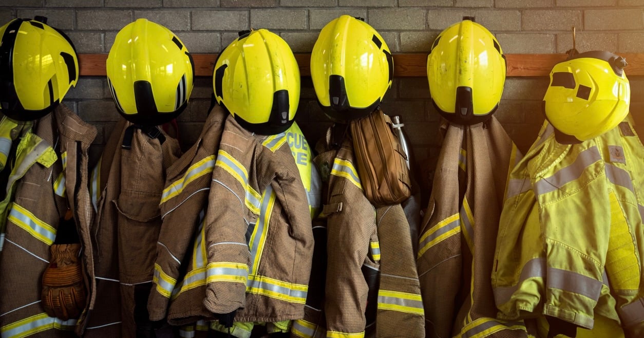 A line of firefighter jackets, helmets, and equipment hanging on a fire department wall.